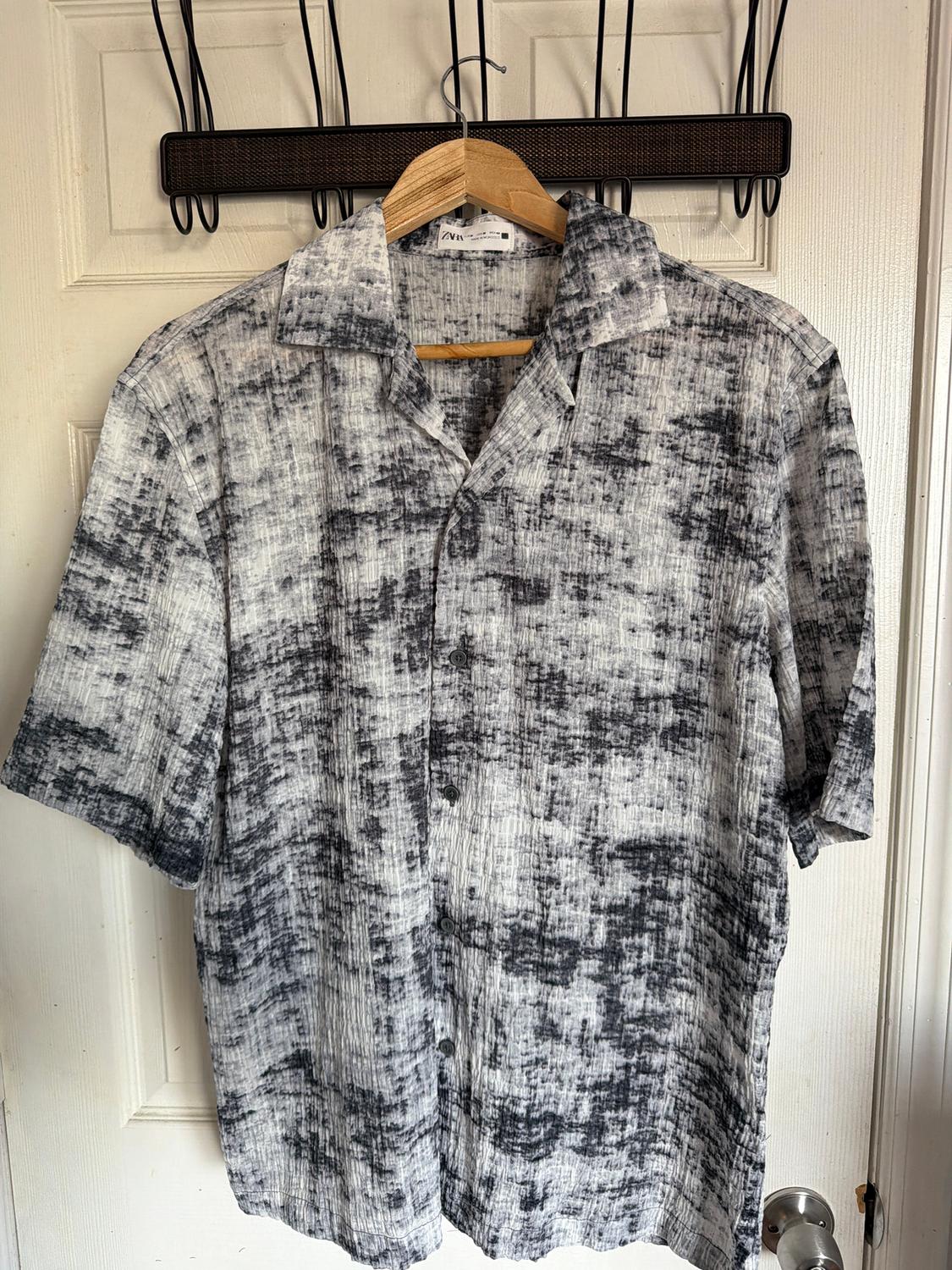 Short-sleeved shirt with black and white abstract print, hanging on a wooden hanger in front of a white door.