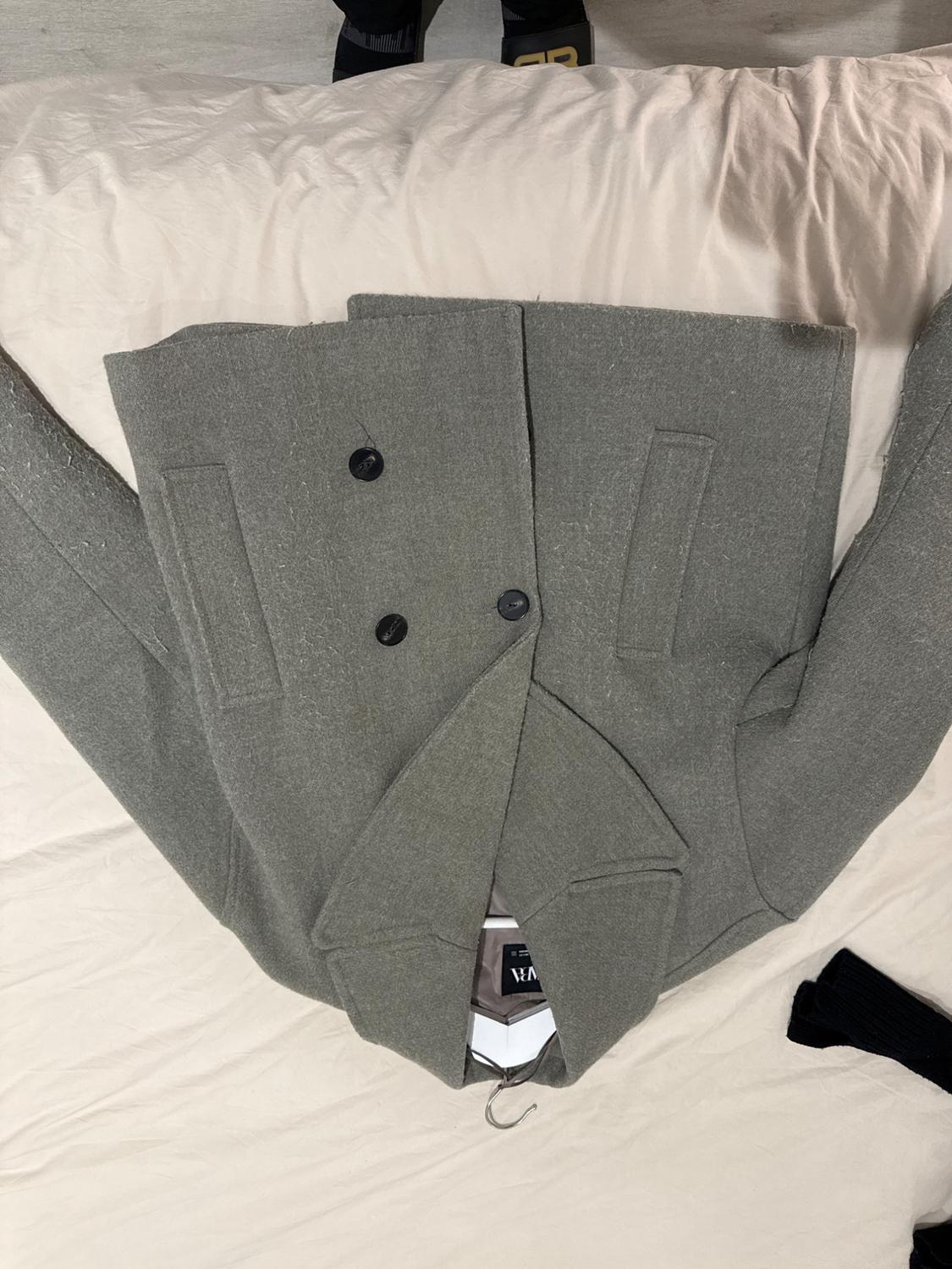 Grey double-breasted wool coat with wide lapels, placed on a beige bed. Part of a pair of black trousers visible.