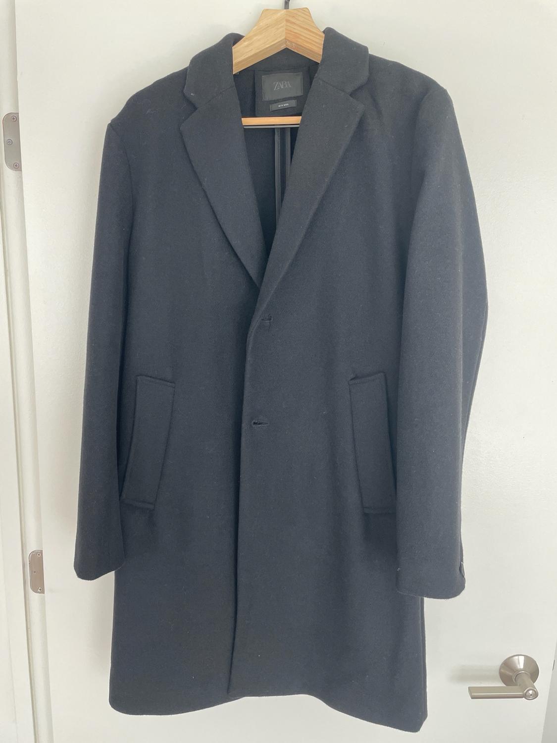 Black coat hanging on a wooden hanger over a white door. The text ZARA is visible on the label.