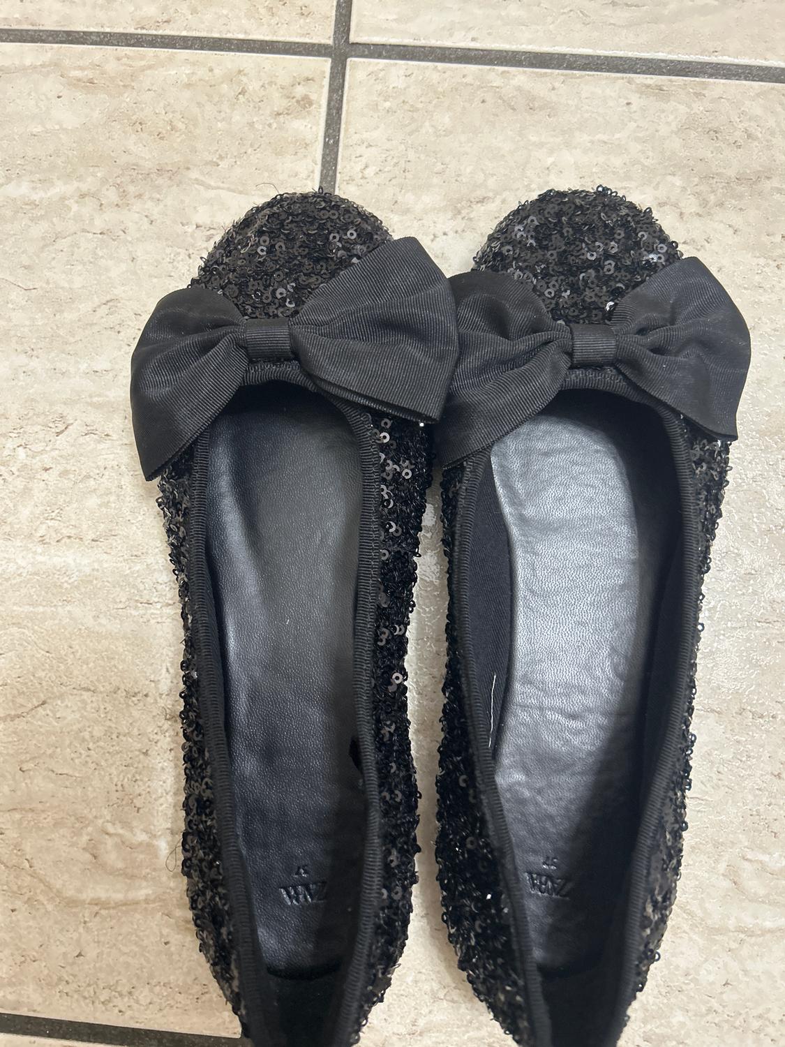 Black sequinned ballet flats with a large bow on top, placed on a beige tiled floor.