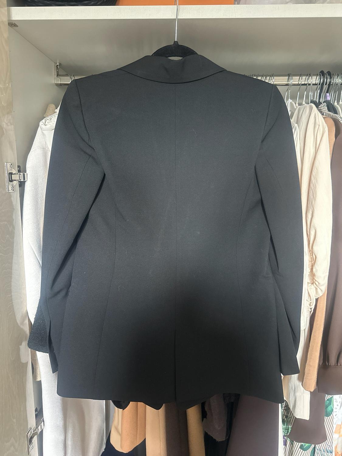 Black blazer seen from behind, hanging in a white wardrobe with other garments in neutral tones in the background. Everyday, relaxed style.