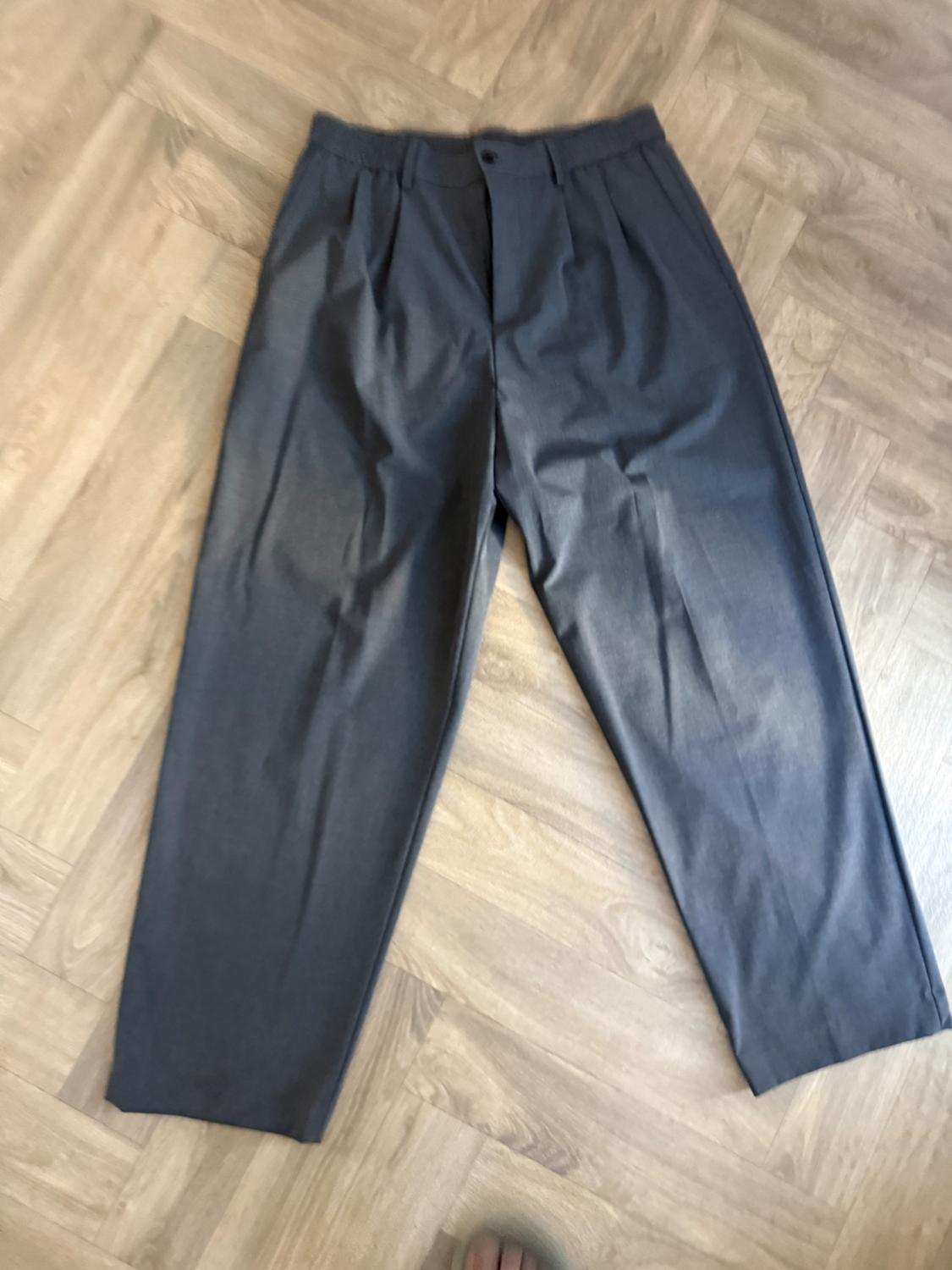 Dark grey pleated wide-leg trousers in a relaxed style, laid out on a light wood floor.