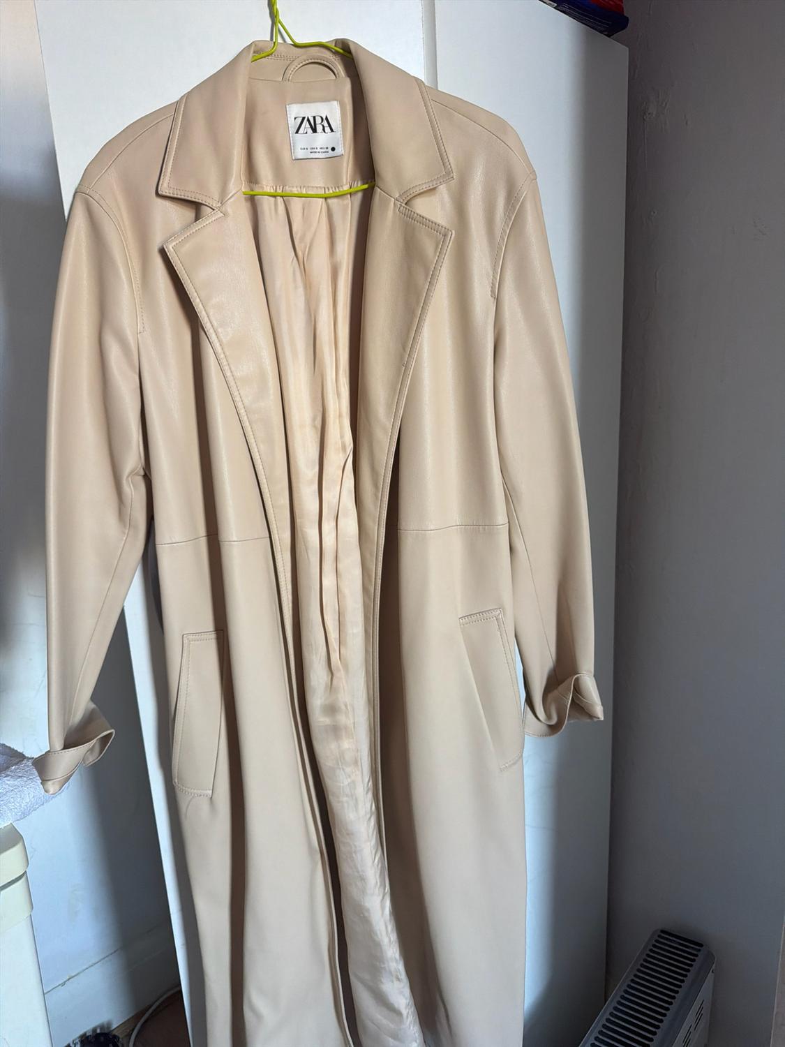 Long beige coat hanging on a hanger in front of a white wardrobe. Interior label with the text ZARA. Home setting.
