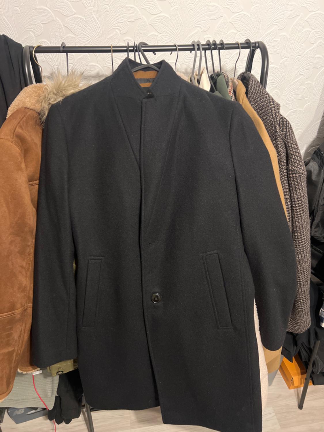 Black coat hanging on a clothes rail next to other garments of various colours and textures. White wallpaper background.