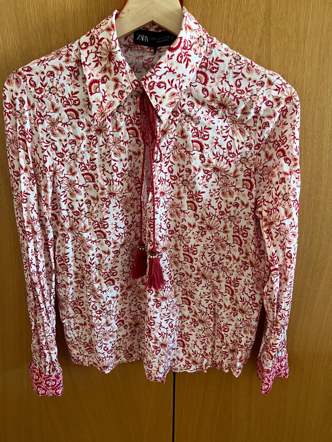 White blouse with a red floral print on a wooden hanger. Featuring a drawstring neckline, red tassels, and the ZARA label.