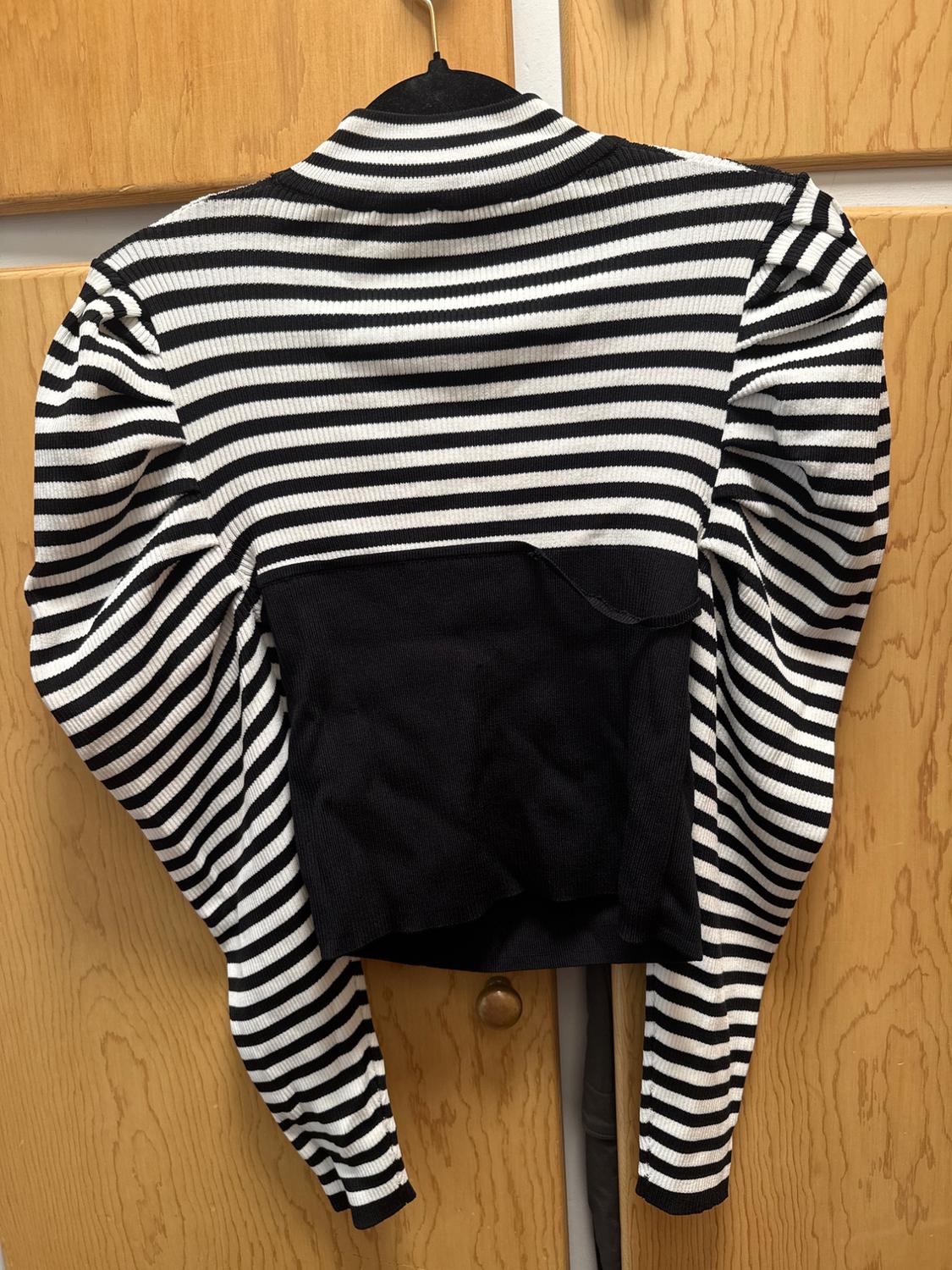 Long-sleeve striped manga top with white and black stripes, puffed shoulders, and a solid black center section, hanging on a coat hanger over a wooden door.