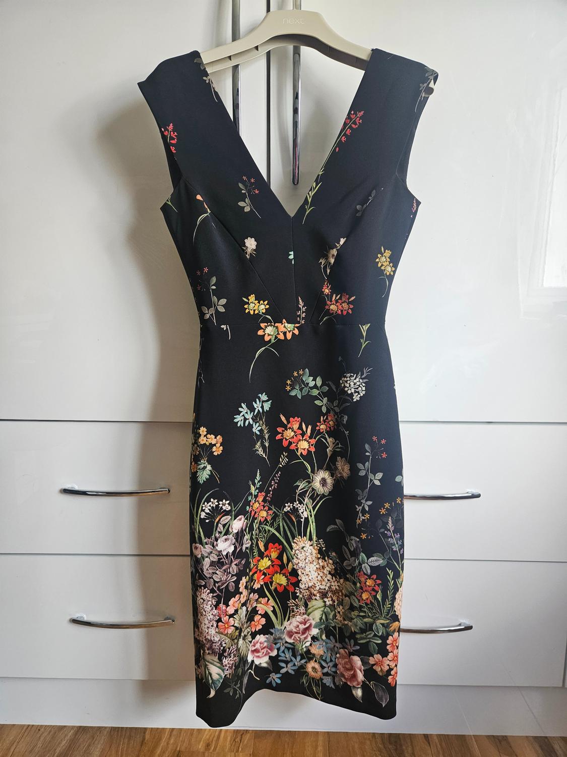 Black dress with floral print and V-neck, hanging on a hanger with the text next in front of a white chest of drawers.