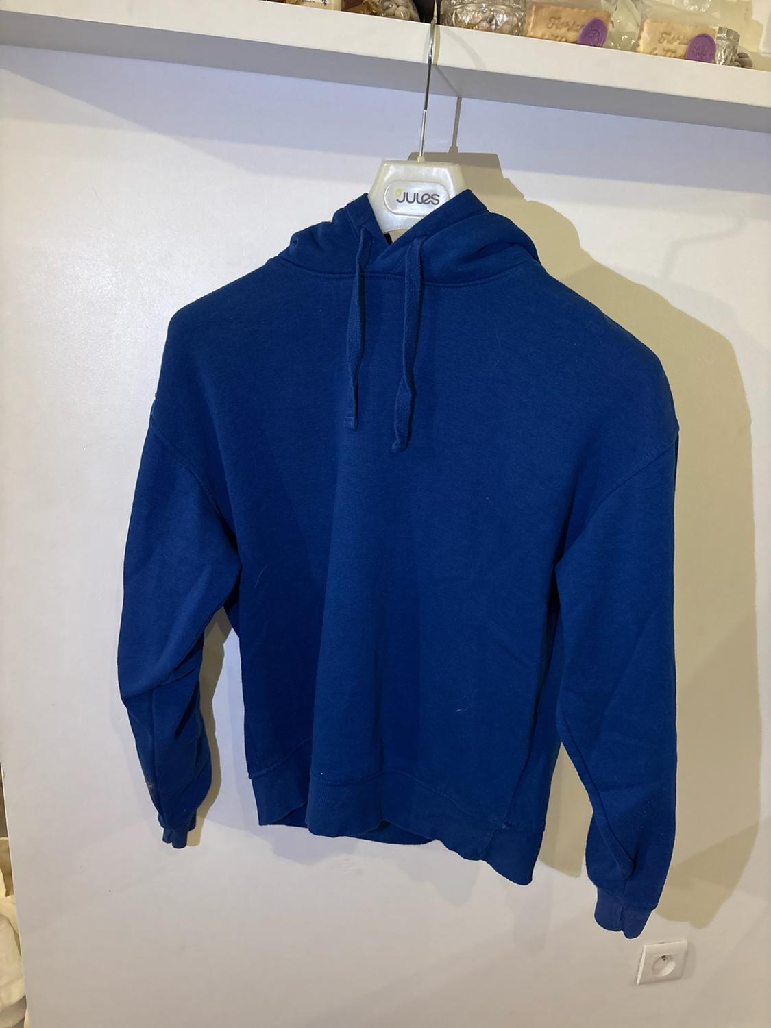 Blue hoodie on a hanger with the text JULES in front of a white wall under a shelf.