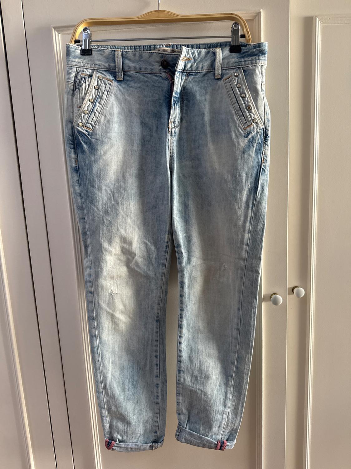 Light blue jeans with stud details on the front pockets, hanging on a hanger against a white wardrobe background.