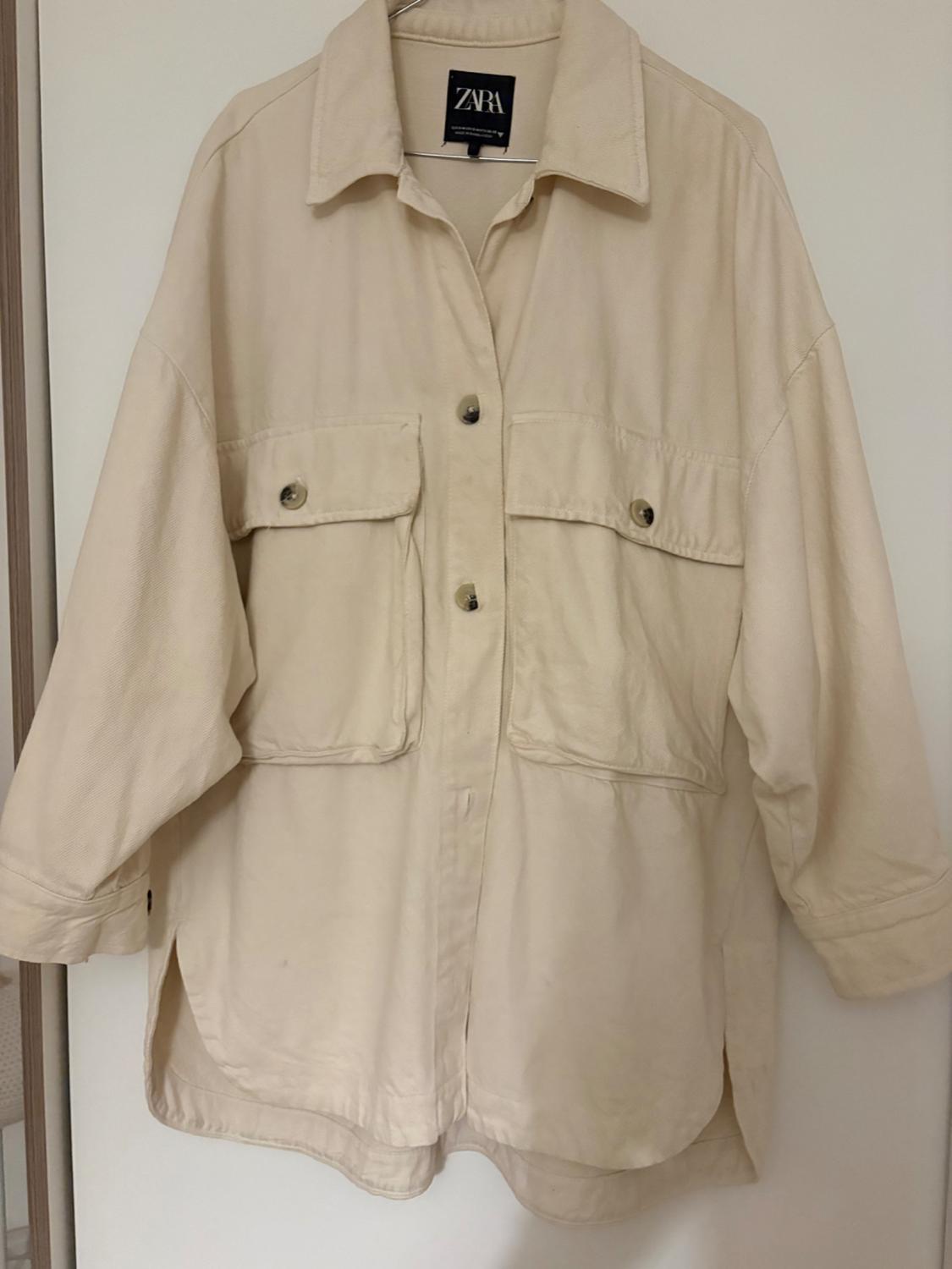 Long-sleeved beige jacket with large pockets and dark buttons, hanging on a hanger. Visible label with the text "ZARA".