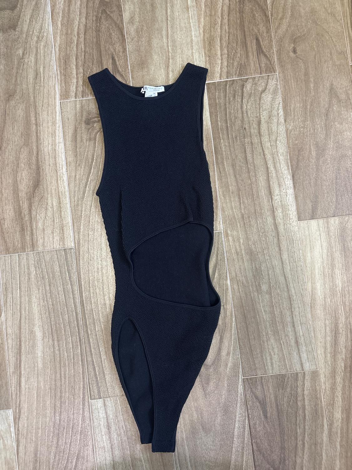 Sleeveless black jumpsuit with an asymmetrical design, placed on a light wooden floor.