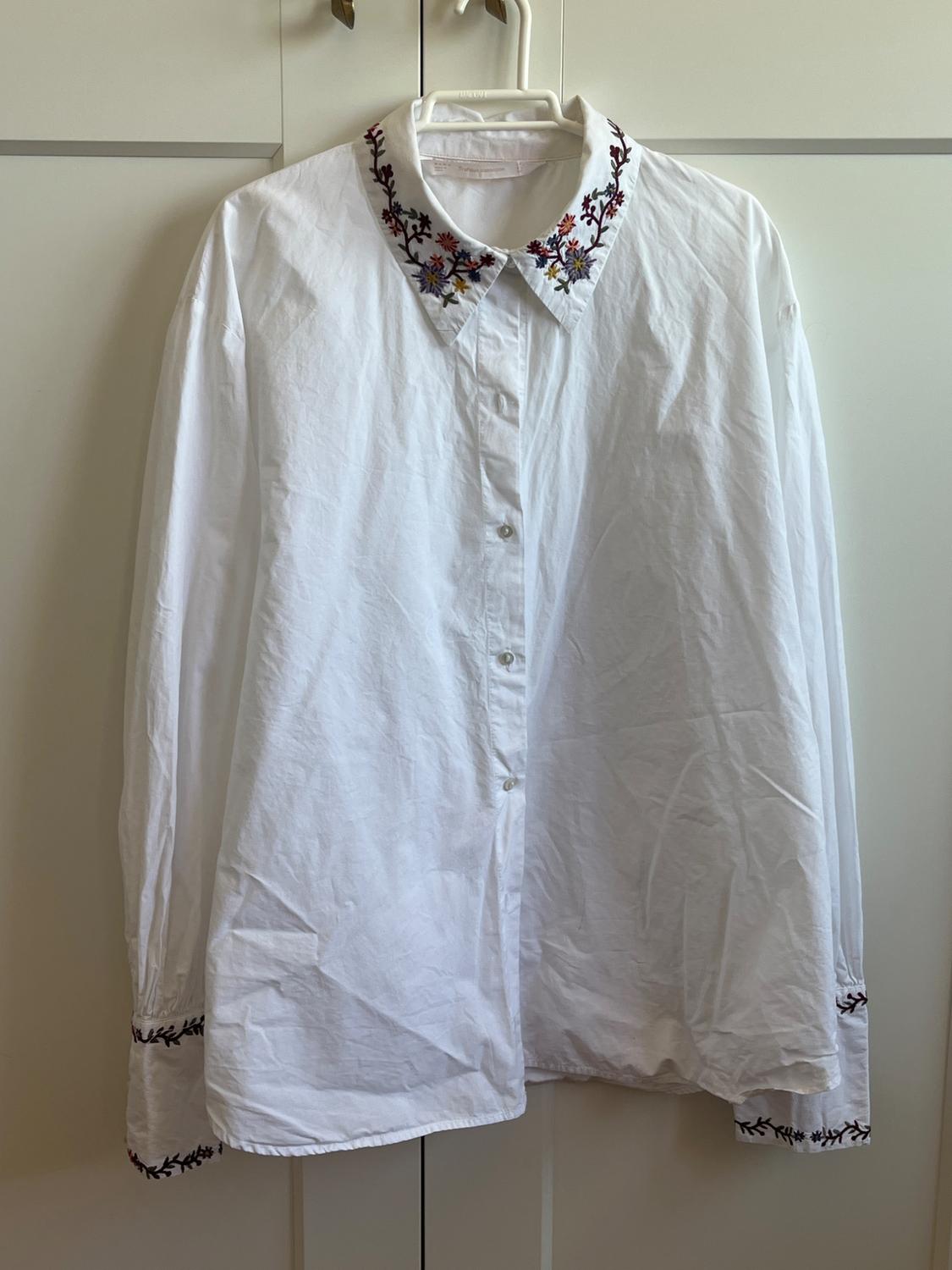 White long-sleeved button-down shirt with floral embroidery on the collar and cuffs, hanging on a hanger in front of a white wardrobe.