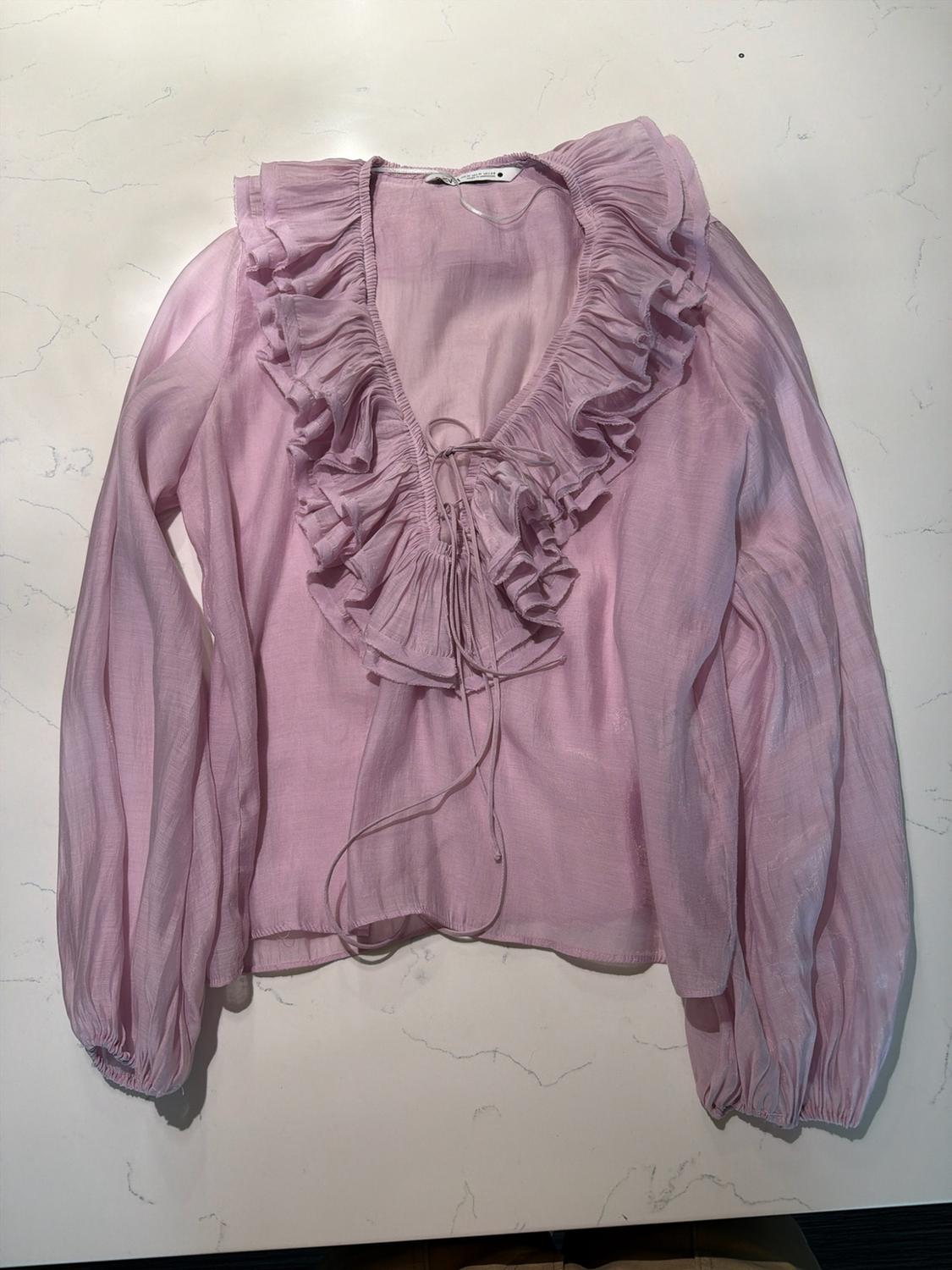 Light pink lightweight blouse, long sleeves, V-neck with large ruffles and front ties, on a white marble surface.