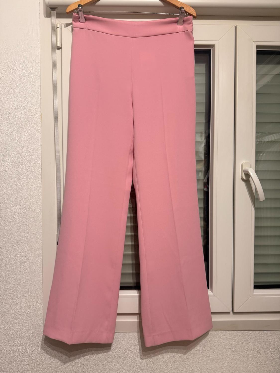 Pale pink wide-leg trousers with a front crease, hanging on a wooden hanger in front of a white window. Casual style.
