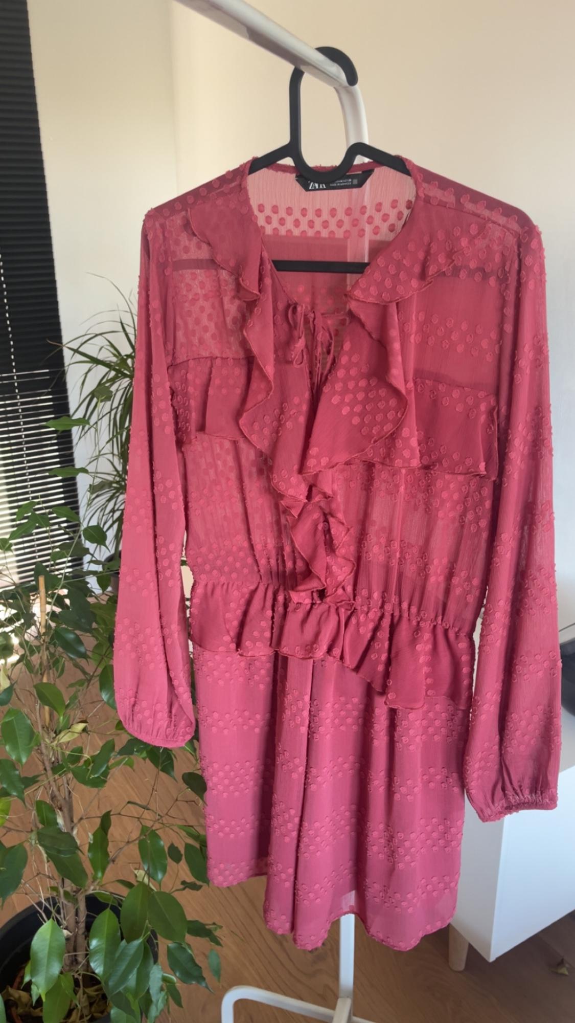 Pink dress with ruffles and polka dot print on a clothes rack. Background with plant and black blind.