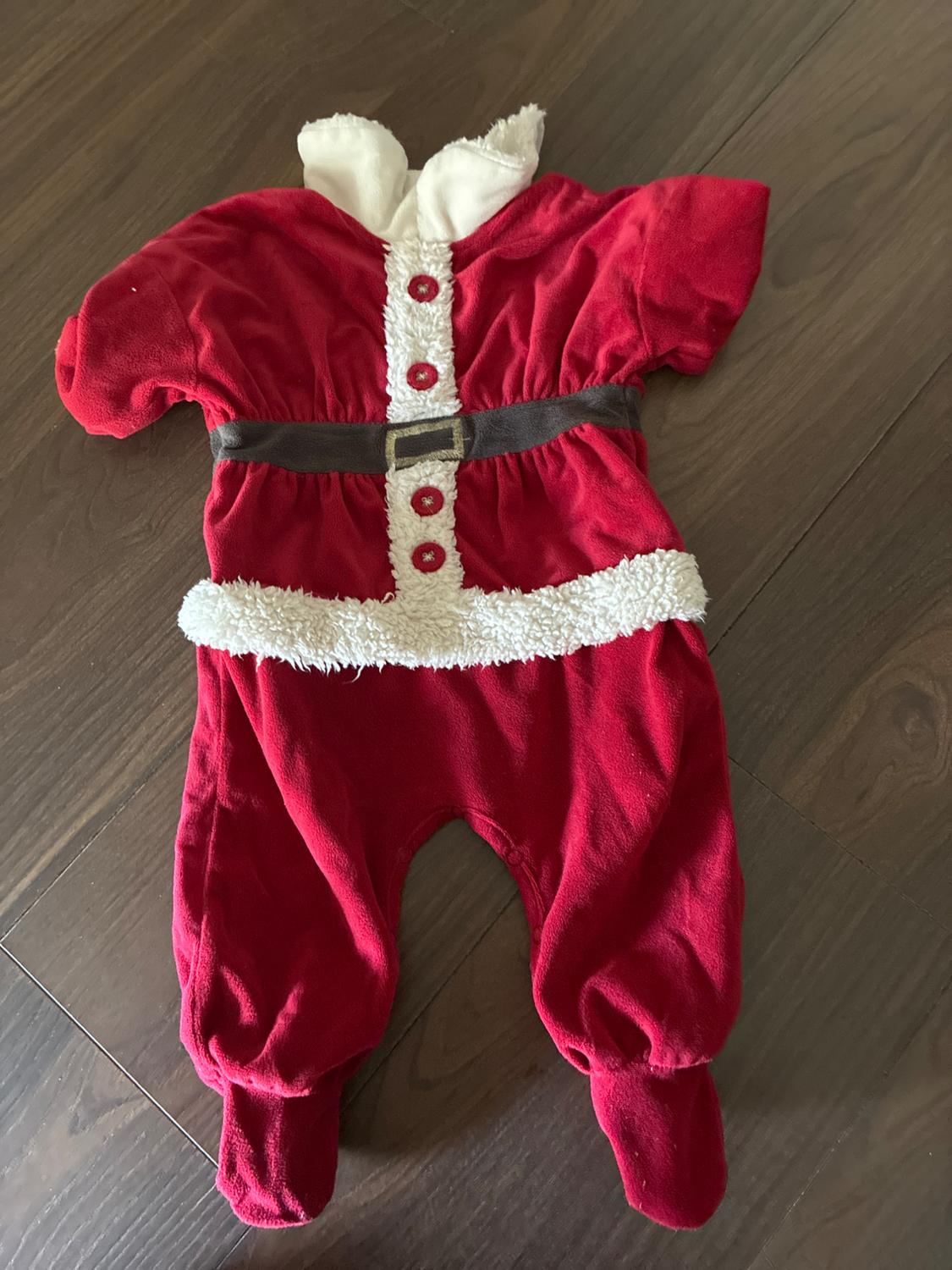 Red Santa-style babygrow with white trim, buttons, and a brown belt, laid out on a wooden floor.