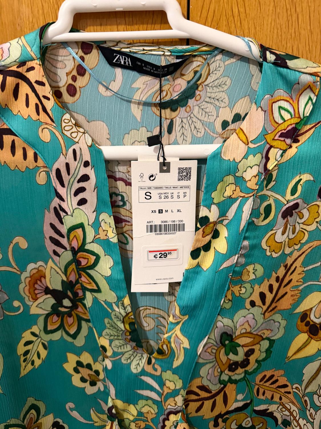 Floral dress in shades of turquoise, yellow, and black hanging on a white hanger. Zara tag with a price of €29.95 visible.
