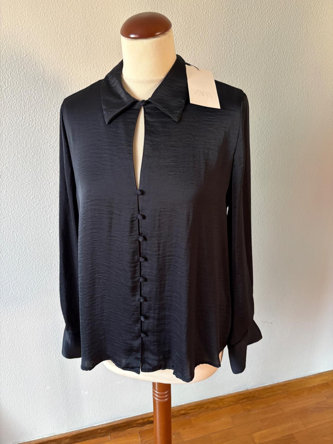 Black long-sleeved collared blouse with an open neckline and buttons on a mannequin. Label with the text ZARA. Light-coloured wall and wooden floor.