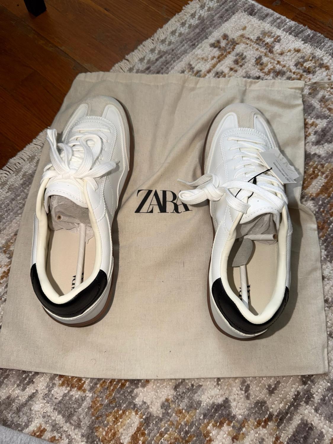 White sneakers with a black heel on a beige fabric bag with the
ZARA text, on a rug and wooden floor.