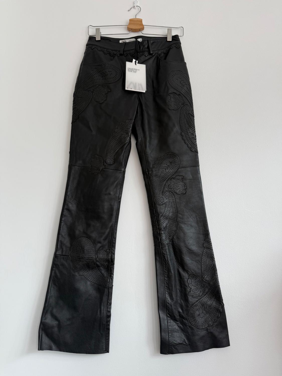 Black trousers hanging on a wooden hanger, with embroidered details and a white label that reads "GRATEFUL HEARTS". White background.