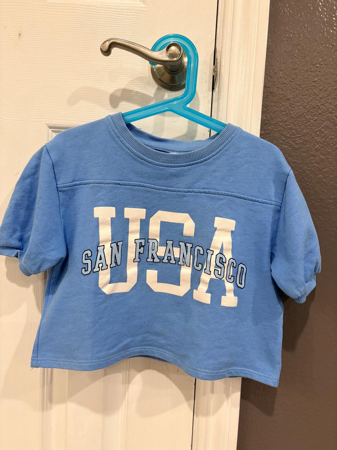 Blue short-sleeved t-shirt hanging on a blue hanger. Text on the front: "USA SAN FRANCISCO". White door background.