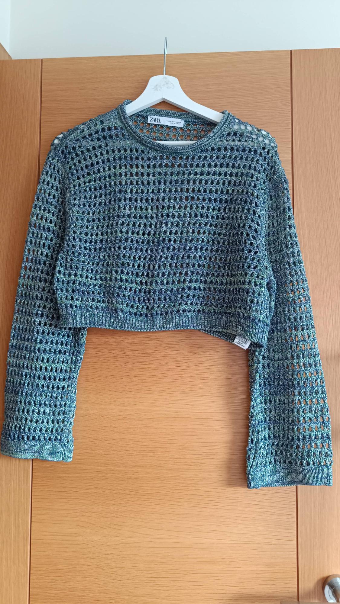 Blue open-knit jumper on a white hanger, hanging on a wooden door. Visible label with text "ZARA".
