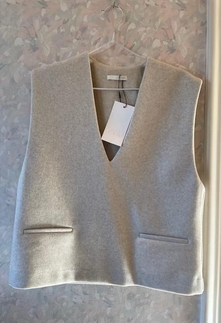 Light grey V-neck waistcoat hanging on a hanger against a floral background. White tag visible in the centre.