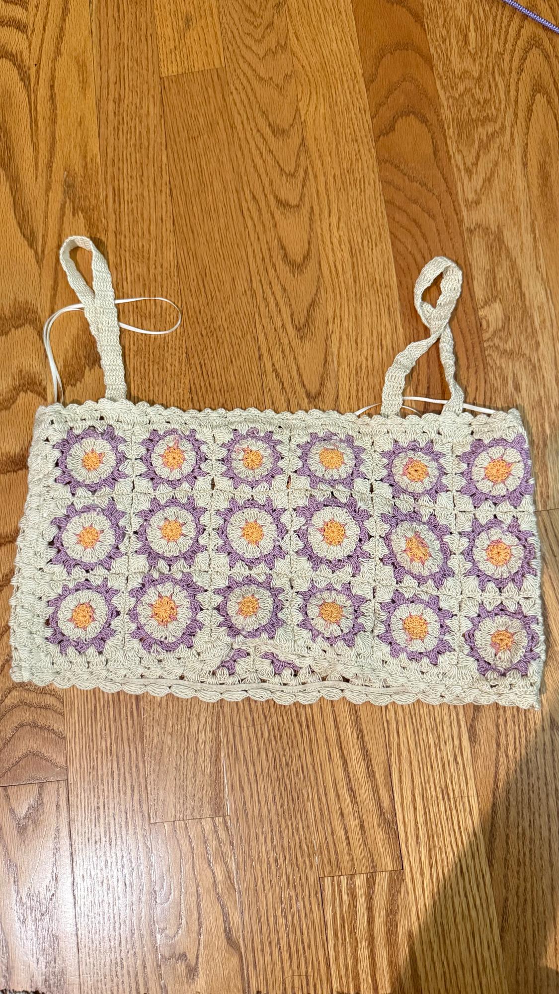 Crochet top with straps on wooden floor. Floral design in shades of cream, lilac, and orange.