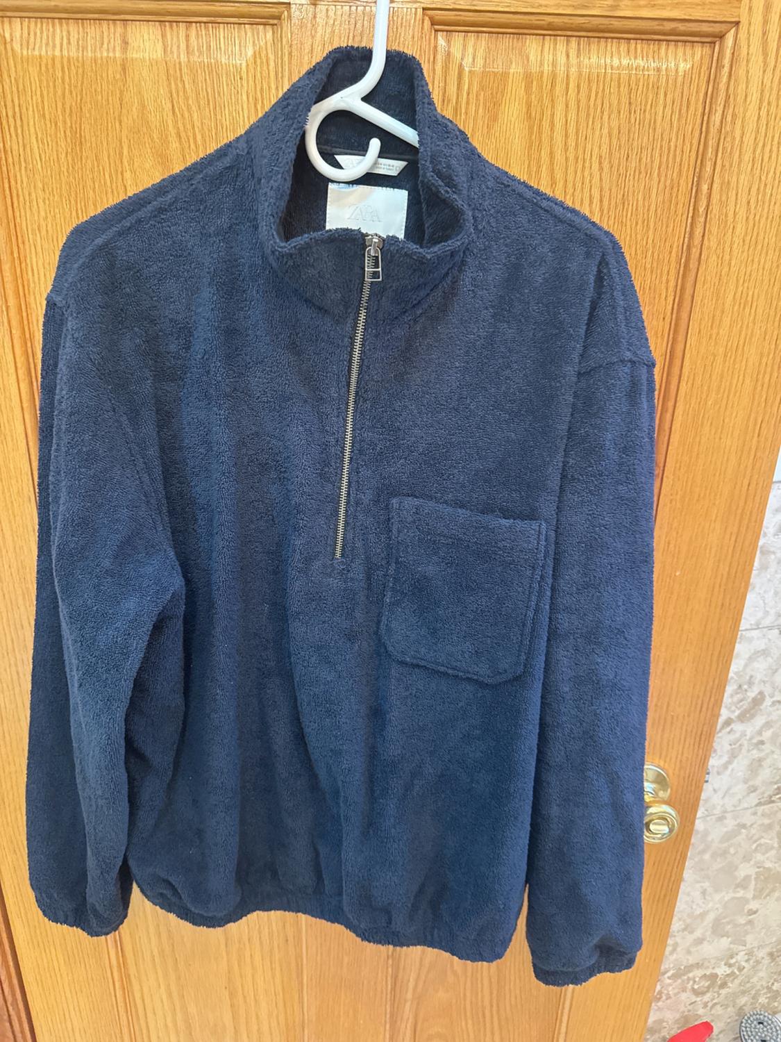 Blue soft fabric jacket with a zip and front pocket, hanging on a white hanger over a wooden door.