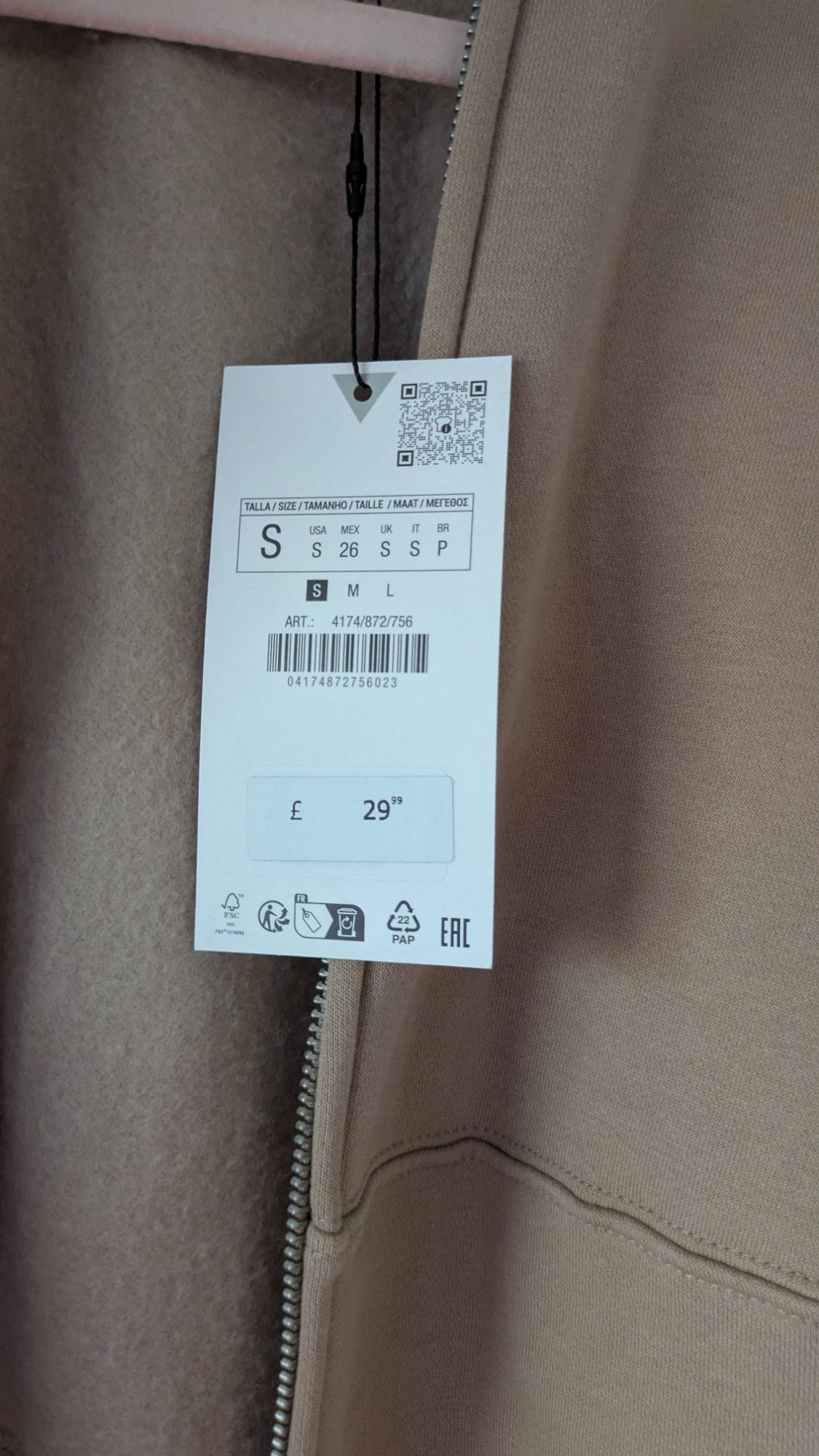 Beige clothing tag with visible zip. Size S, price £29.99. Barcode and recycling symbols on tag.