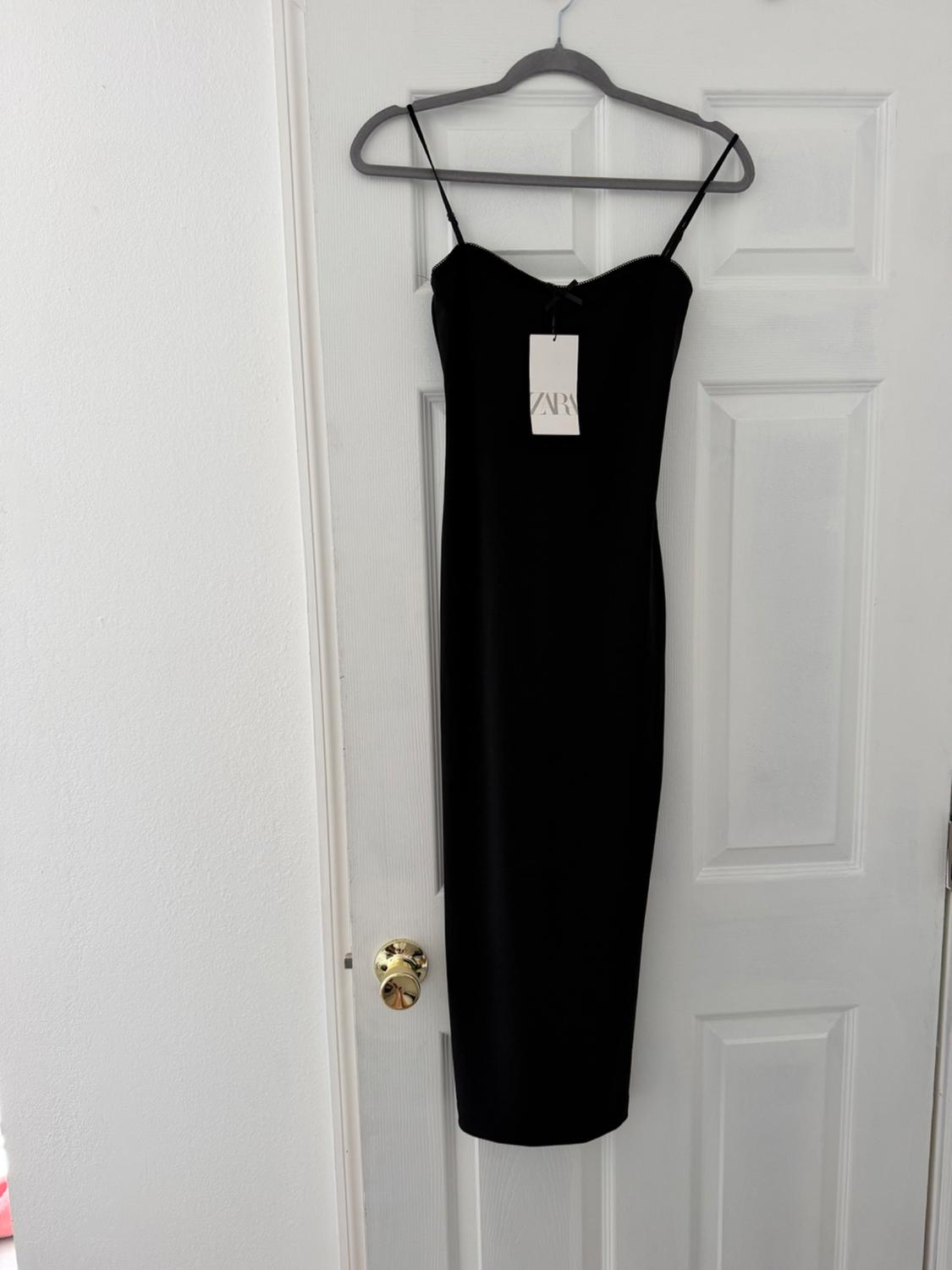 Black slip dress hanging on a hanger over a white door. Visible tag with the text "ZARA".
