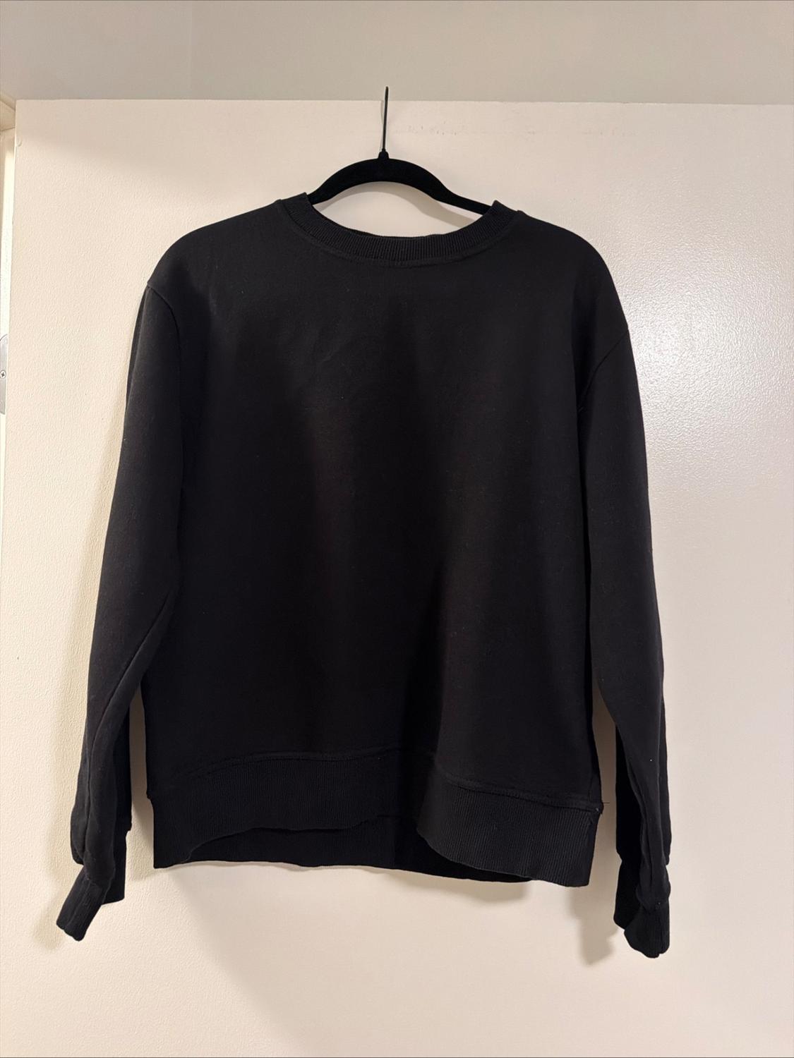 Basic black long-sleeved, round-neck sweatshirt, hanging on a black hanger against a white wall background.