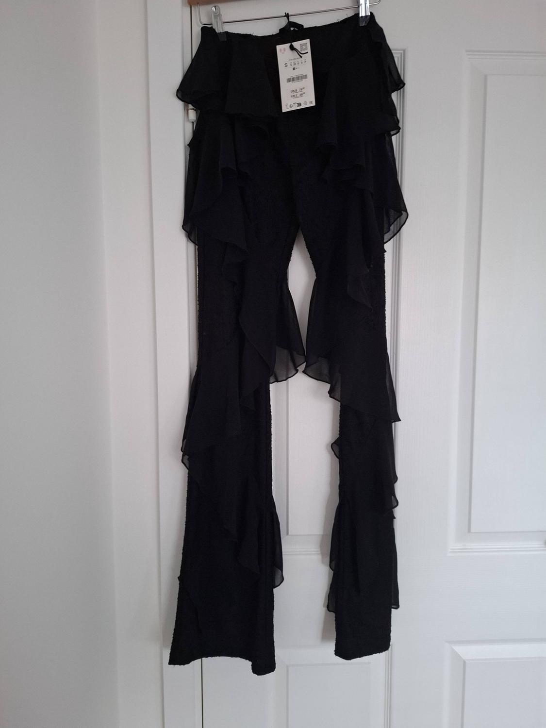 Black trousers with semi-transparent ruffles on the legs, hanging from a hanger on a white door.
