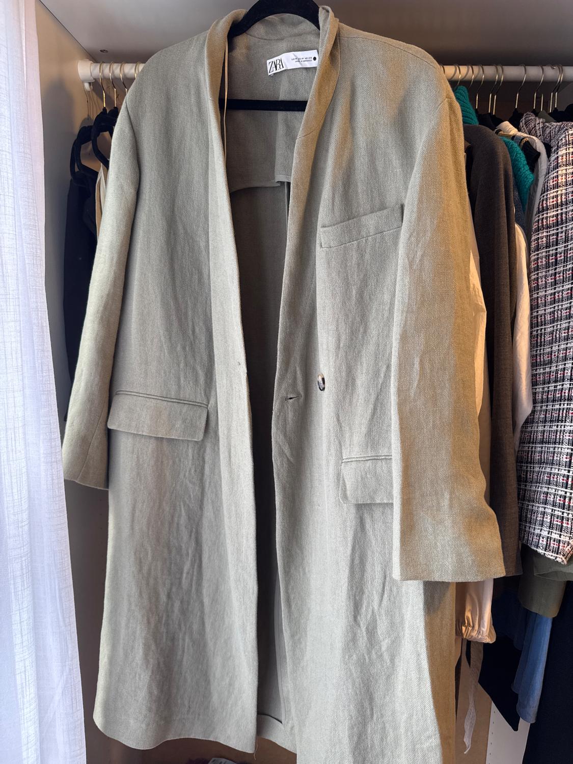 Light grey long coat hanging on a hanger inside a wardrobe with other clothes. The label on the neck reads ZARA.