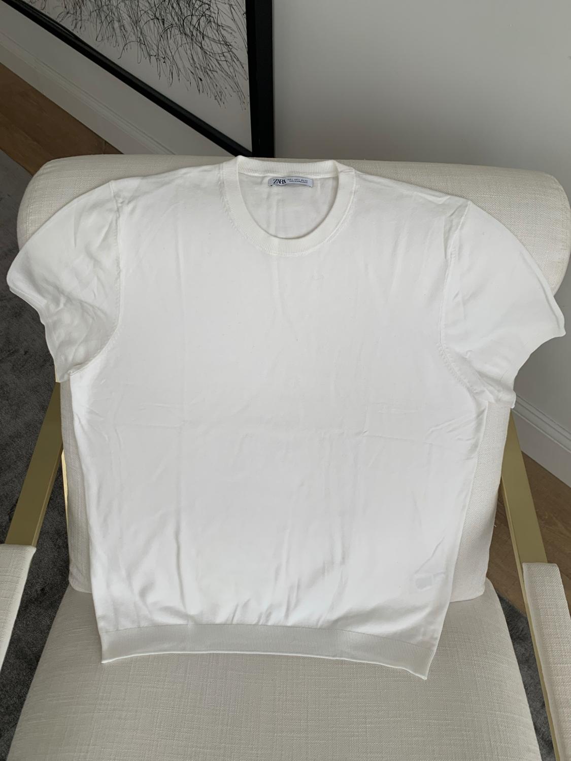 White short-sleeved T-shirt with a ZARA label on a light-coloured armchair with gold-coloured arms. In the background, part of a framed picture.