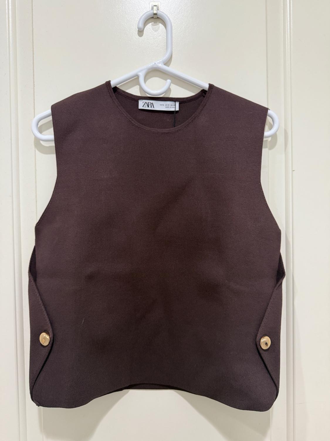 Sleeveless brown vest hanging on a white hanger, with decorative buttons on the sides. Visible label: ZARA.