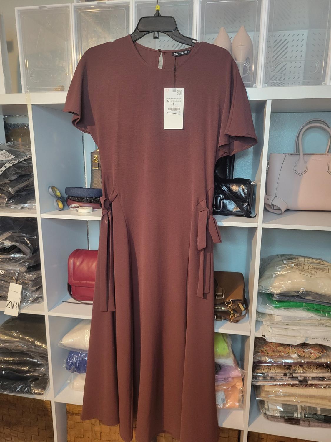 Brown dress with short sleeves on hanger, visible tag. White shelf background with purses and folded clothes.