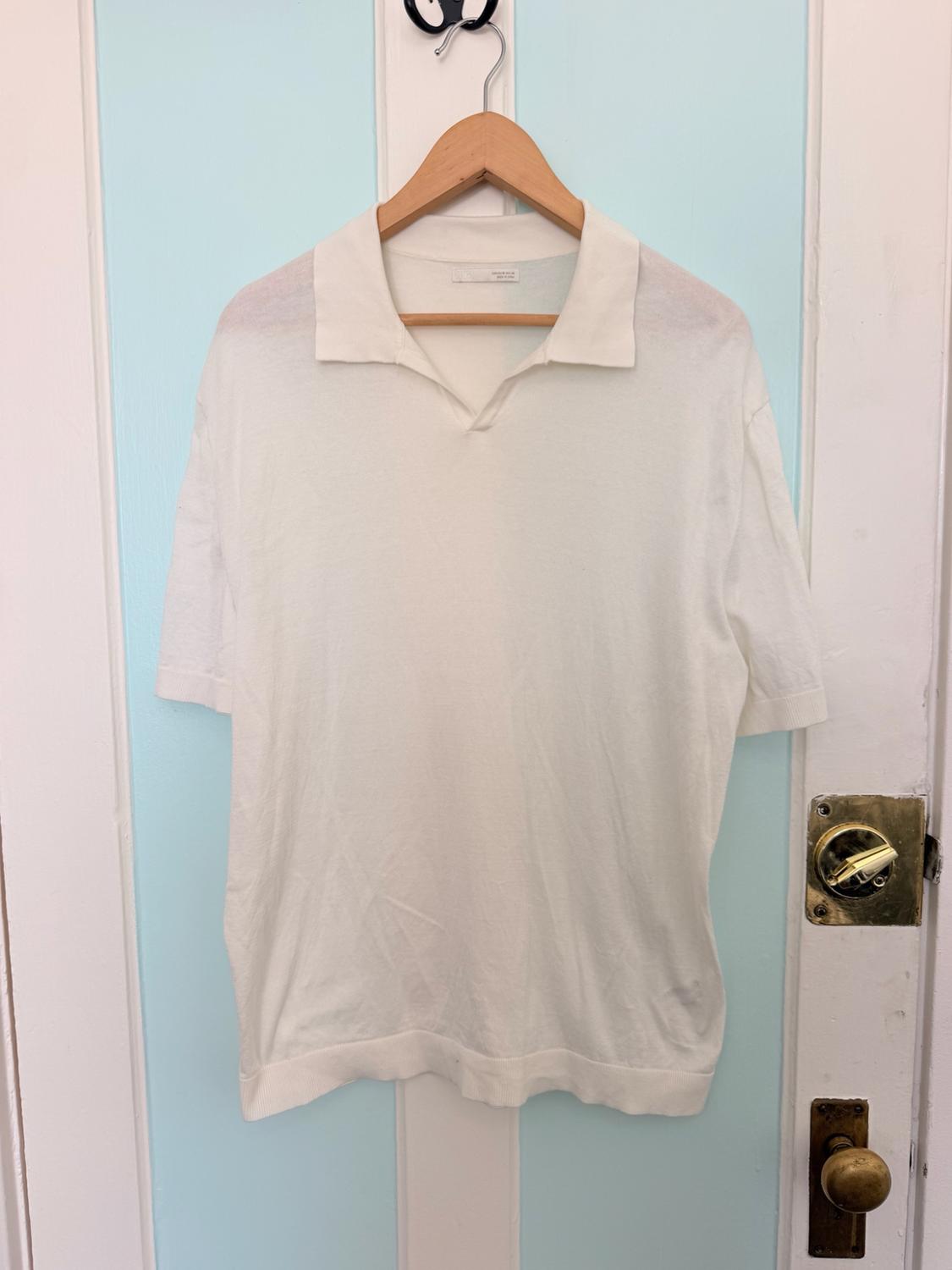 White short-sleeved polo shirt on a wooden hanger, hanging on a white and light blue striped door with a gold handle. Relaxed style.