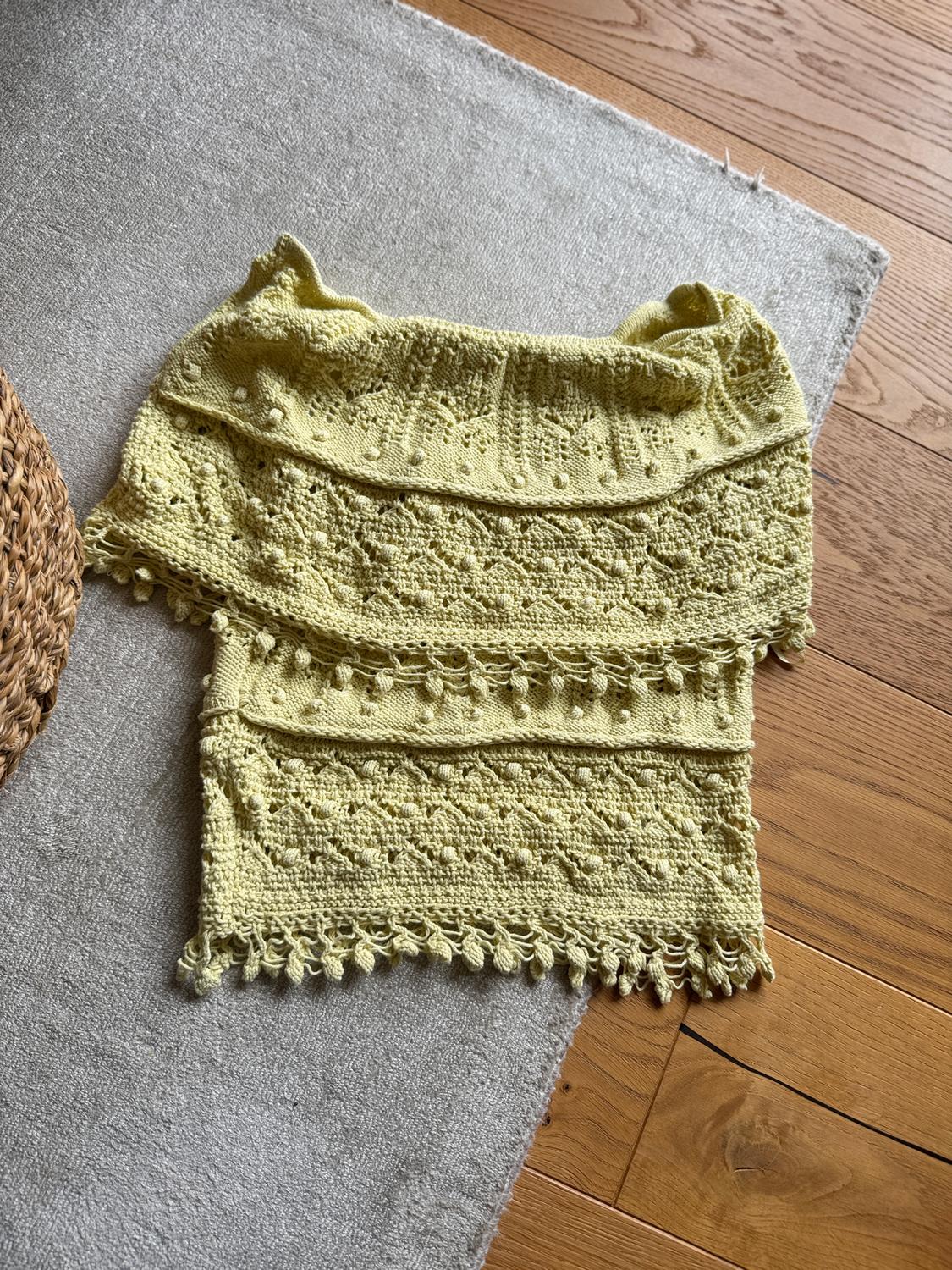 Yellow knitted shawl with stitch details and tassels, on a grey rug and light wooden floor. Cosy and warm atmosphere.