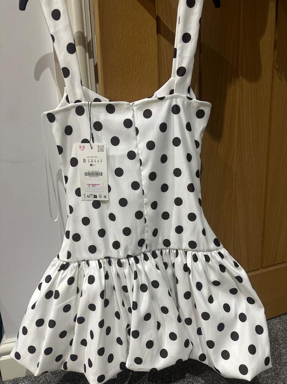 White dress with black polka dots and wide straps, hanging on a wooden door. Visible tag with a price of €25.