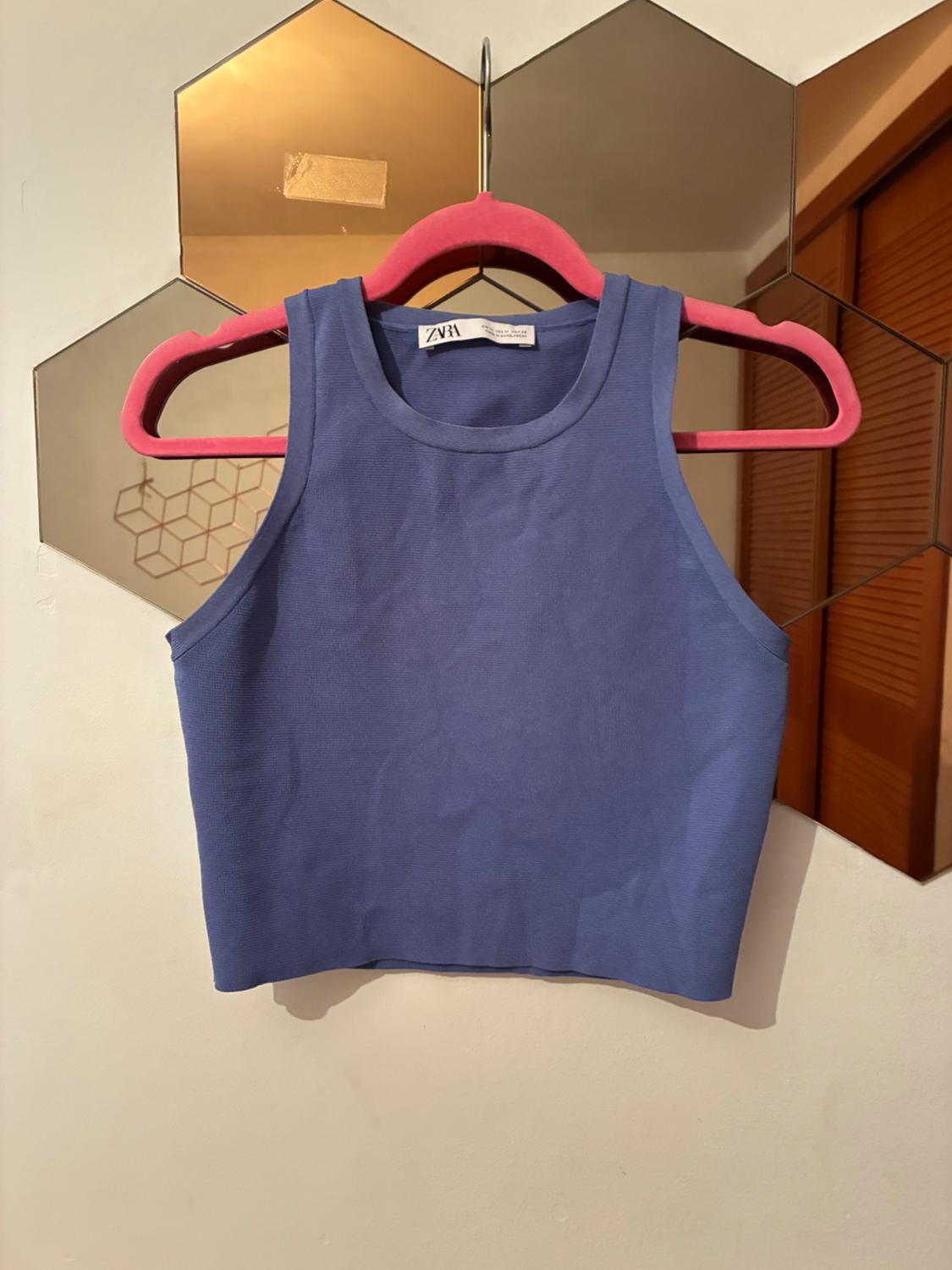 Blue sleeveless crop top with ZARA tag hanging on a pink hanger in front of a hexagonal panel mirror on a white wall.