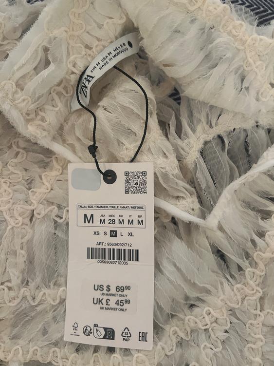 White clothing label with fringed texture, shows size M and price: US $69.90, UK £45.99. Made in Morocco. Barcode visible.