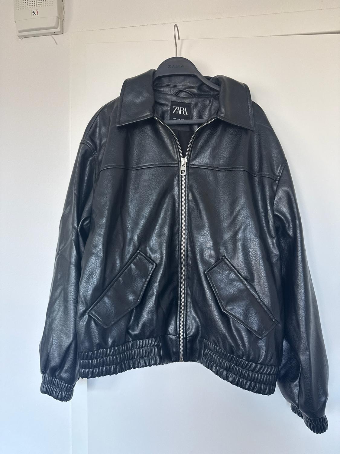 Black jacket with a zipper, collar, pockets, and elastic hem, hanging on a hanger over a white door. Visible text: ZARA.