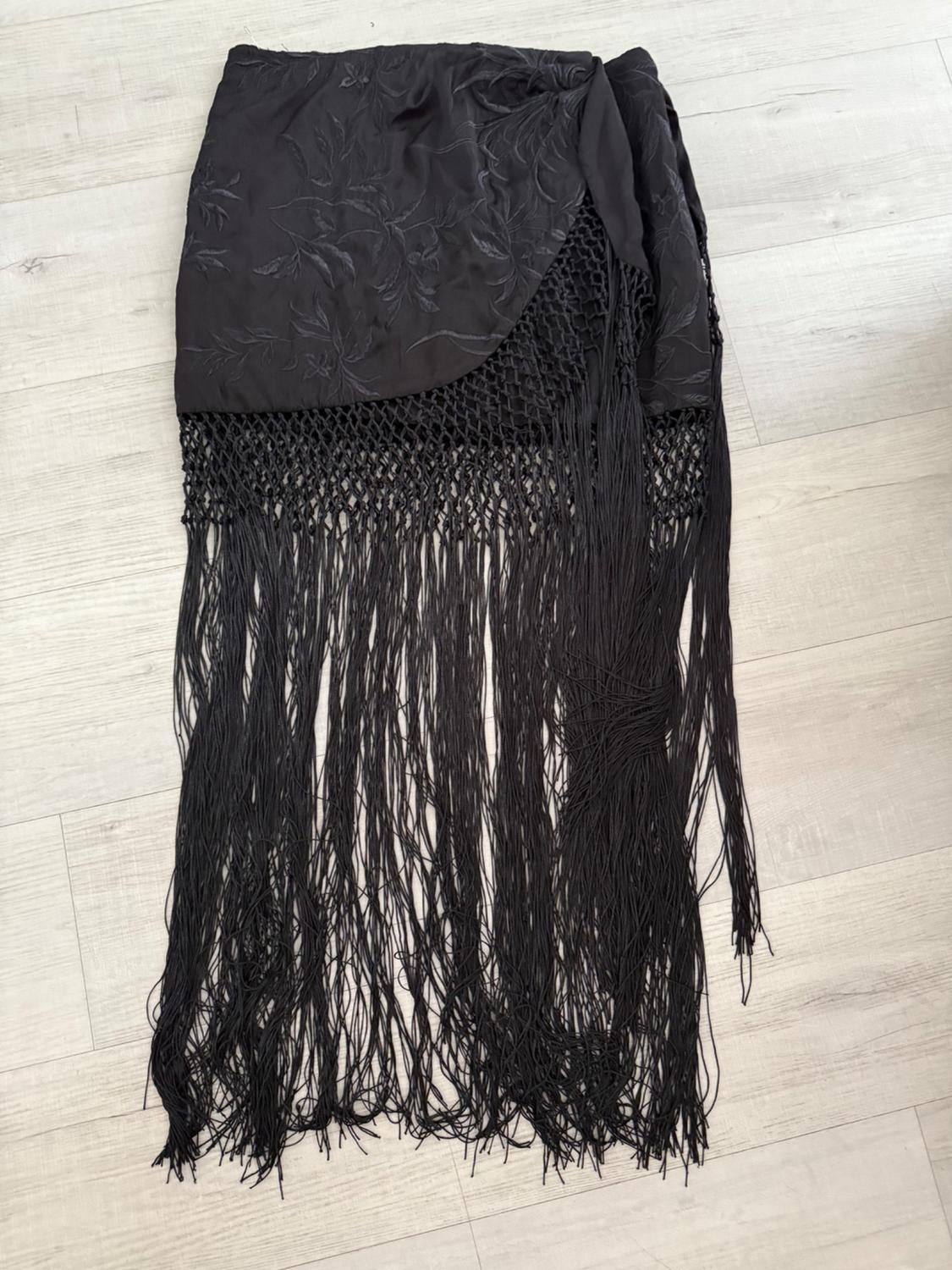 Black skirt with floral embroidery on the upper part, net fabric detail and long fringes, on a light wood floor.