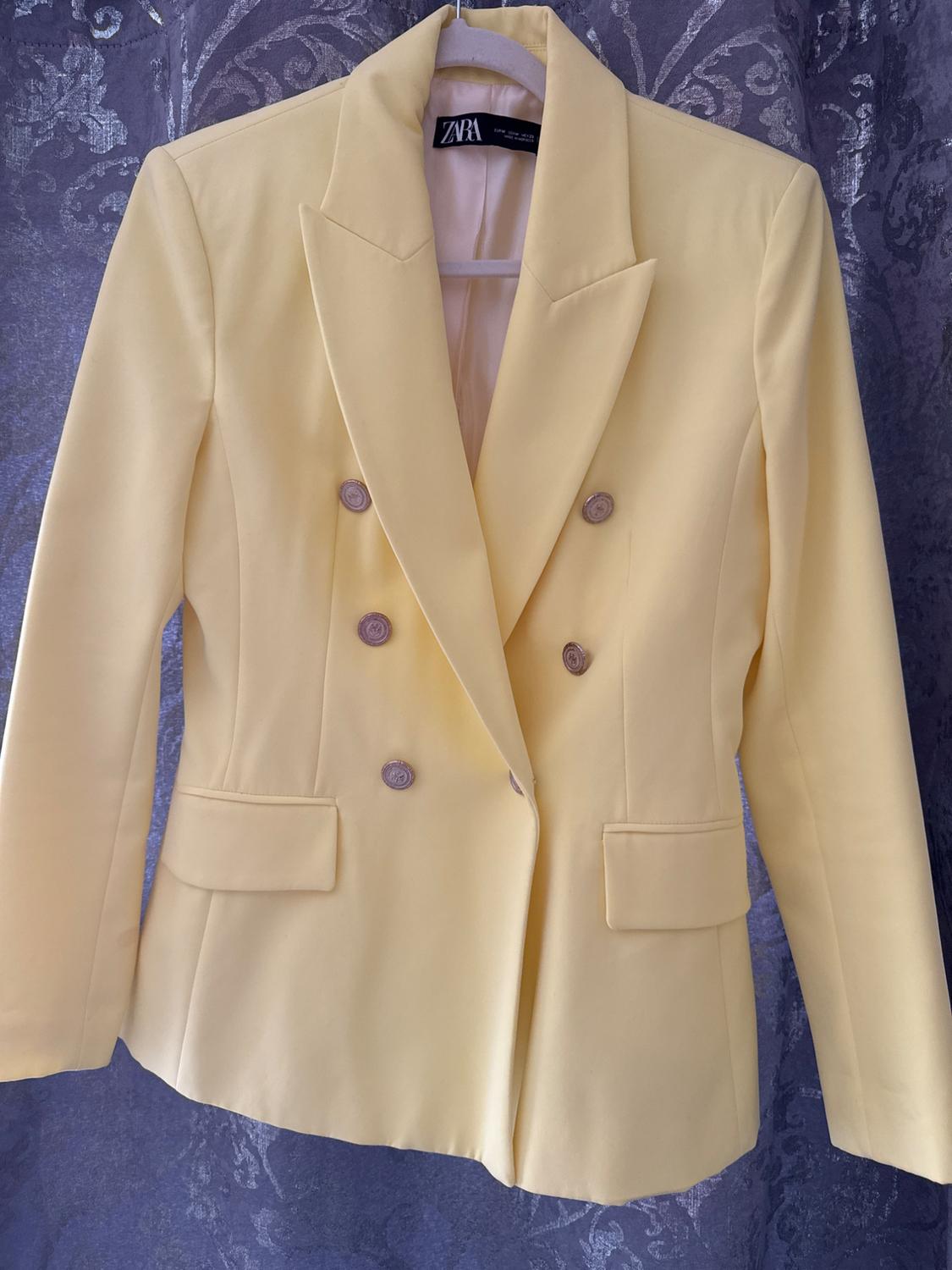 Yellow double-breasted jacket hanging on a hanger, with a gray floral-patterned curtain in the background. Visible tag: ZARA.