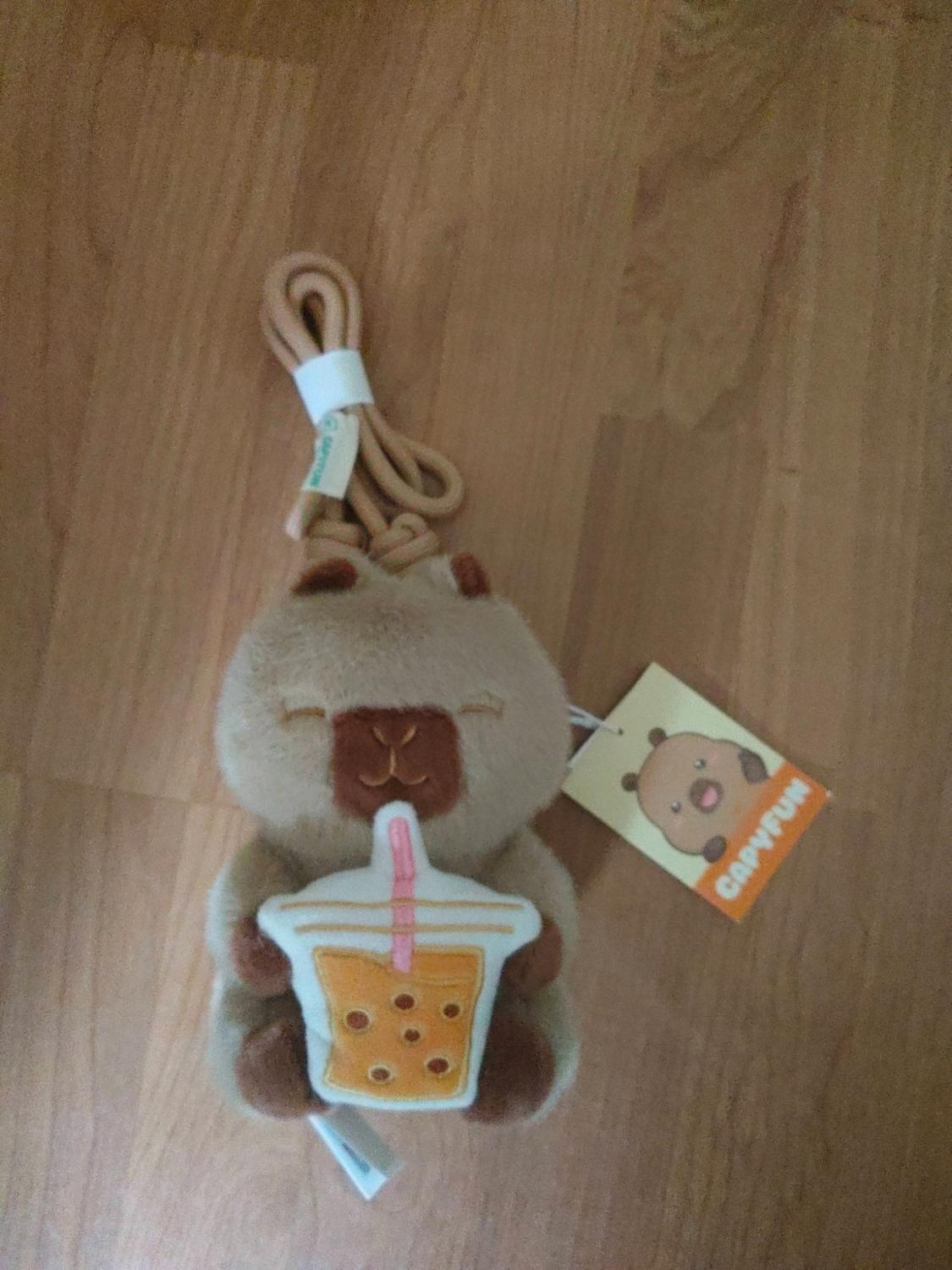 Capybara plush toy with cord holding a bubble tea. Includes a tag bearing the text CAPYFUN, on a wooden floor.
