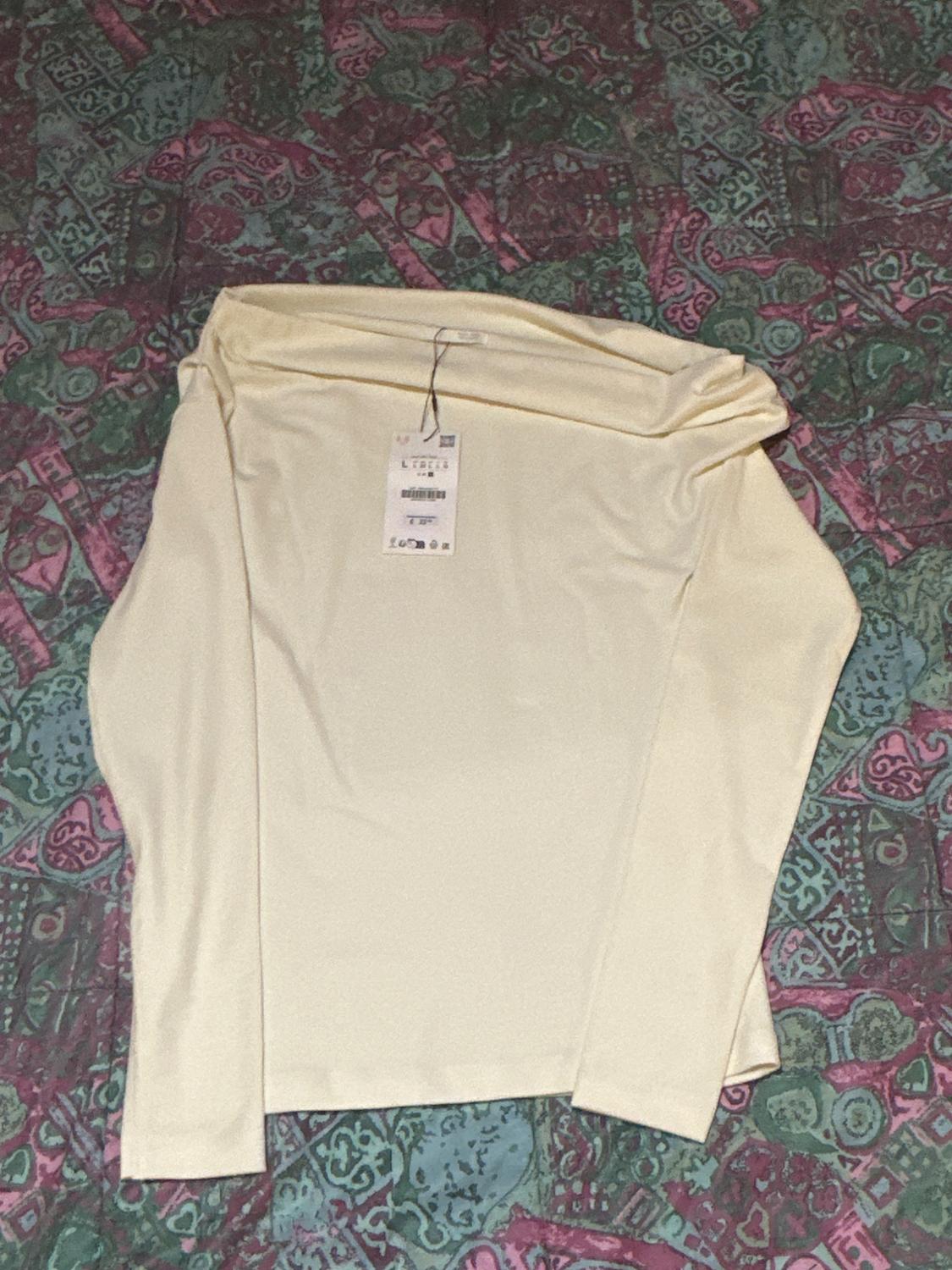 Cream long-sleeved T-shirt with swing tag, on a bedspread with a geometric pattern in shades of green and pink.