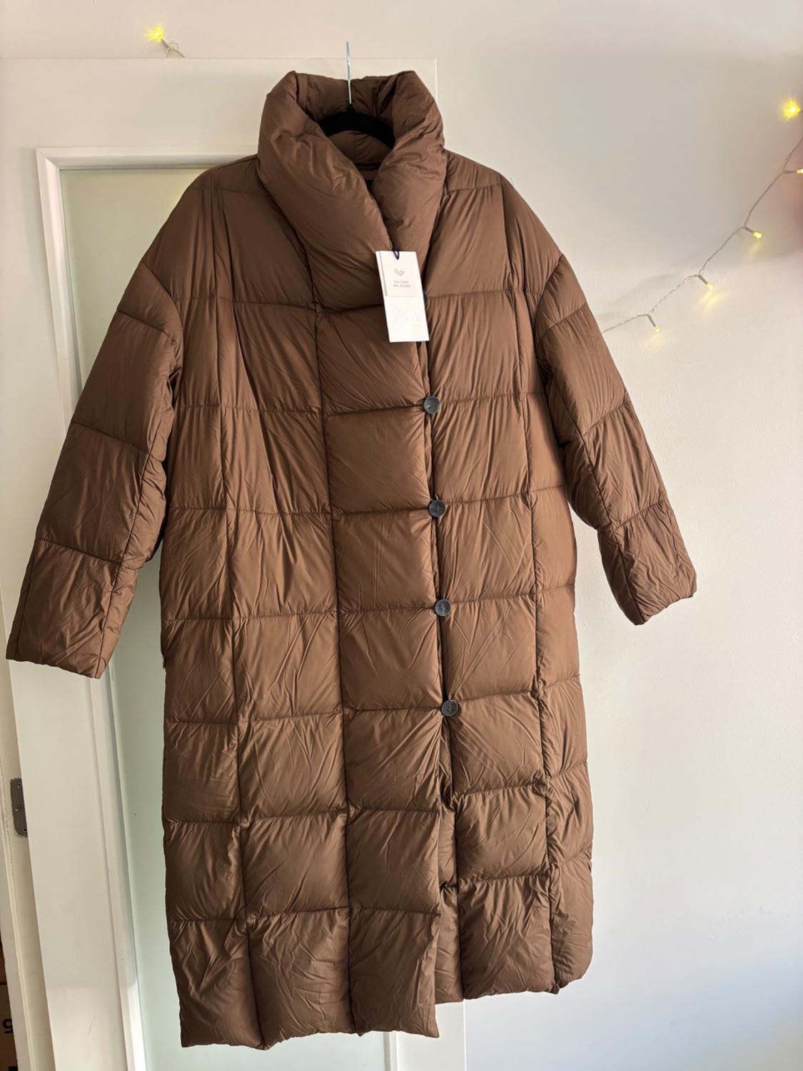 Brown padded coat hanging on a white door, with visible tag. Decorative lights in the background.