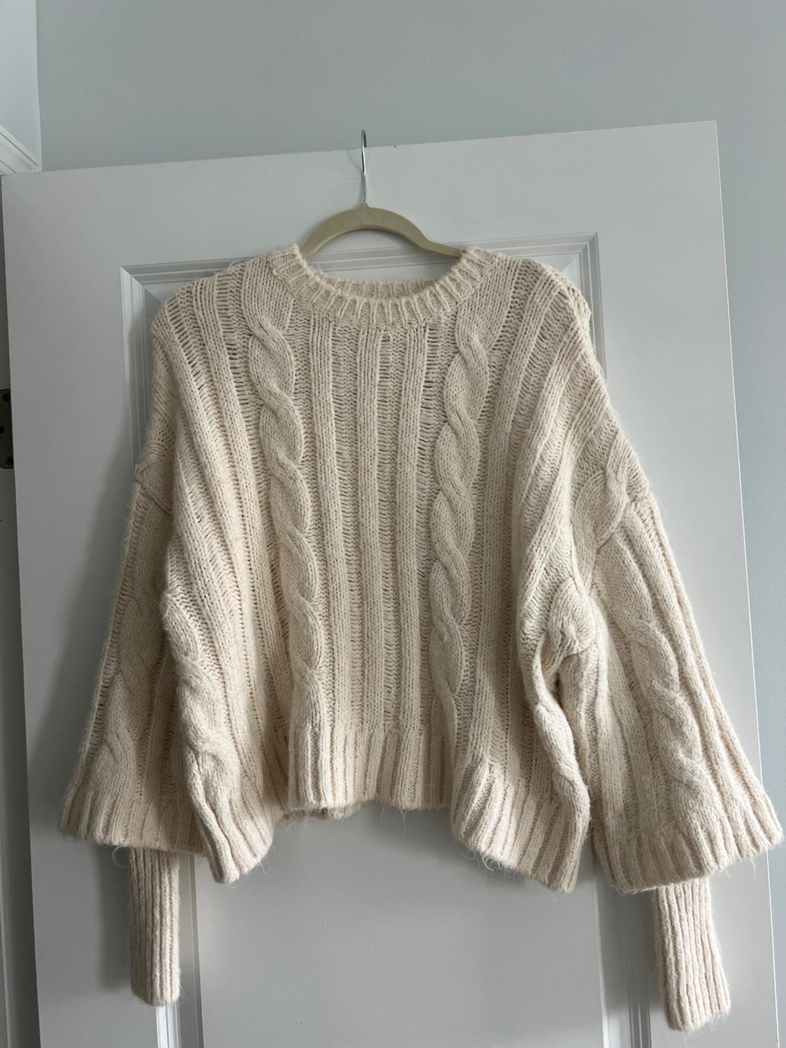 Chunky knit cream-colored sweater with a cable knit design hanging on a hanger over a white door.