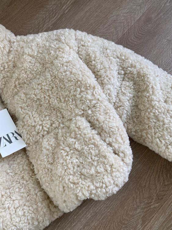 Cream-colored fluffy textured jacket on a wooden floor. Visible label with "ZARA" text in the bottom left corner.